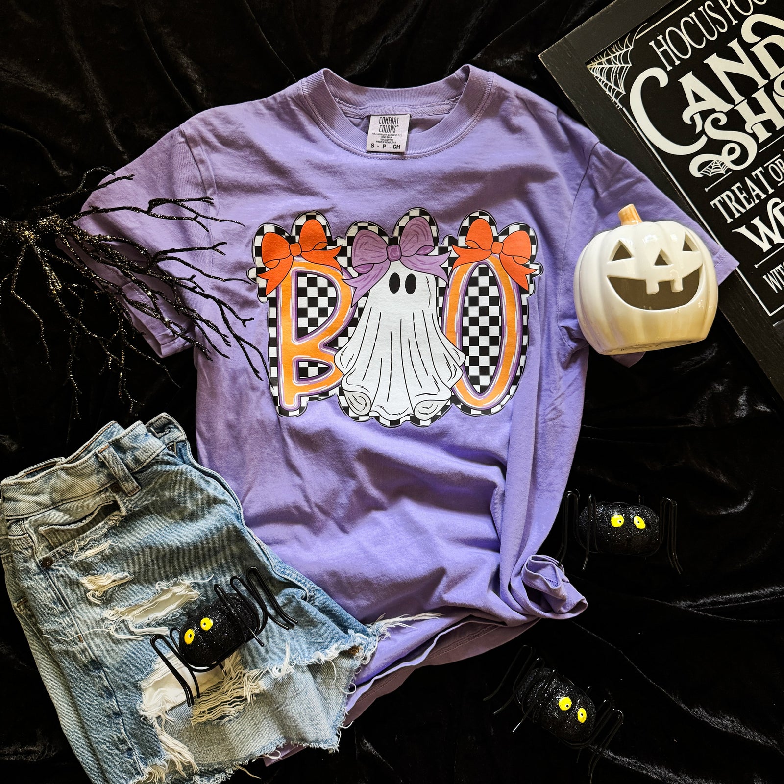 Purple t-shirt with Halloween-themed design featuring ghosts and checkered letters, paired with denim shorts and a small pumpkin on a dark background.