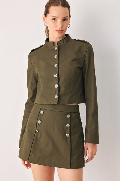 A woman modeling a matching olive green cropped jacket and high-waisted mini skirt set with silver-tone buttons, photographed against a plain light gray studio background.