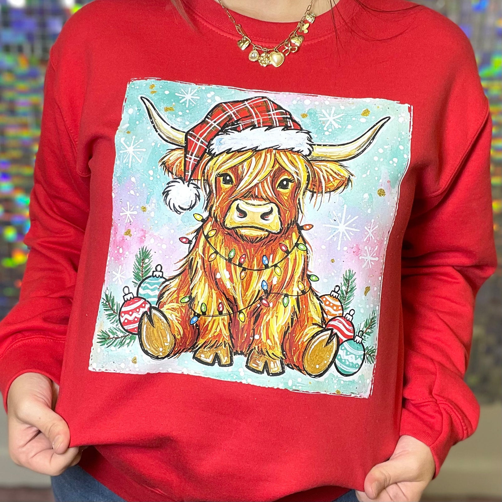 Red sweatshirt with a festive cow design featuring a Santa hat and Christmas ornaments on a colorful background.