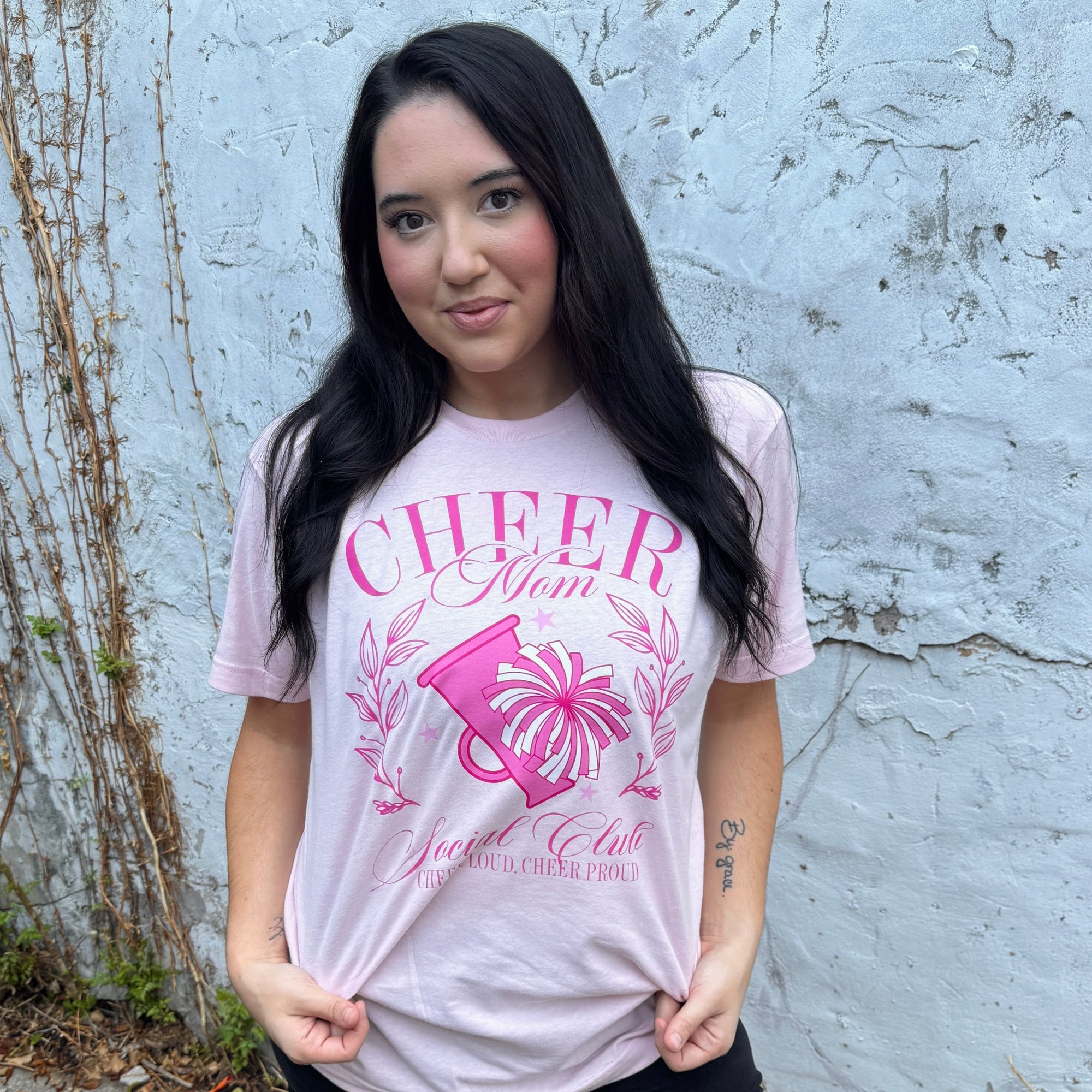 Woman wearing a pink 'Cheer Mom' t-shirt against a textured wall.