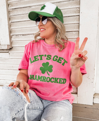 Person wearing a pink t-shirt with 'Let's Get Hamrocked' text and a green cap, sitting against a wooden wall.