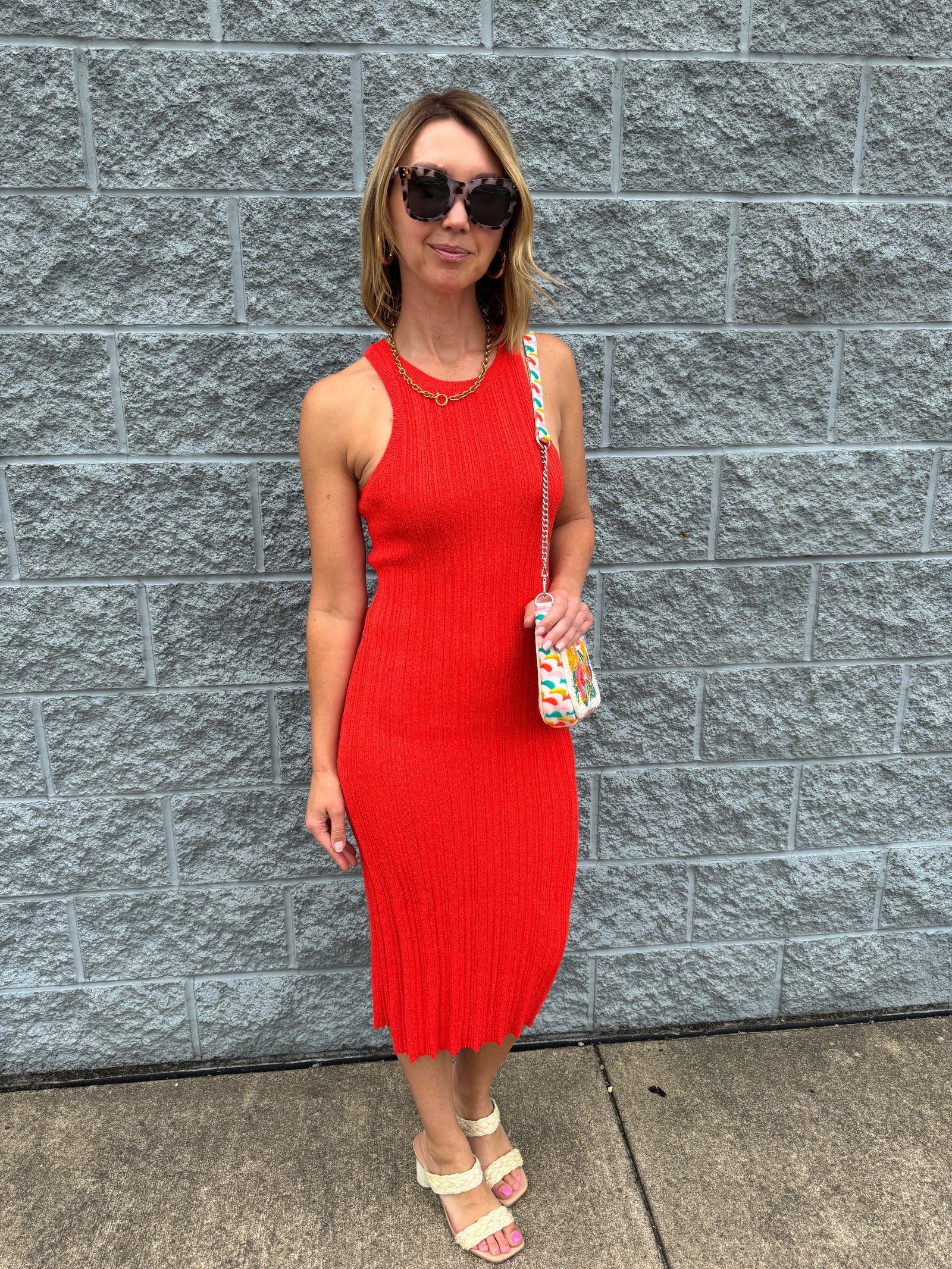 A person wearing a red sleeveless knee-length dress with a pleated design, paired with white sandals and carrying a small multicolored bag.