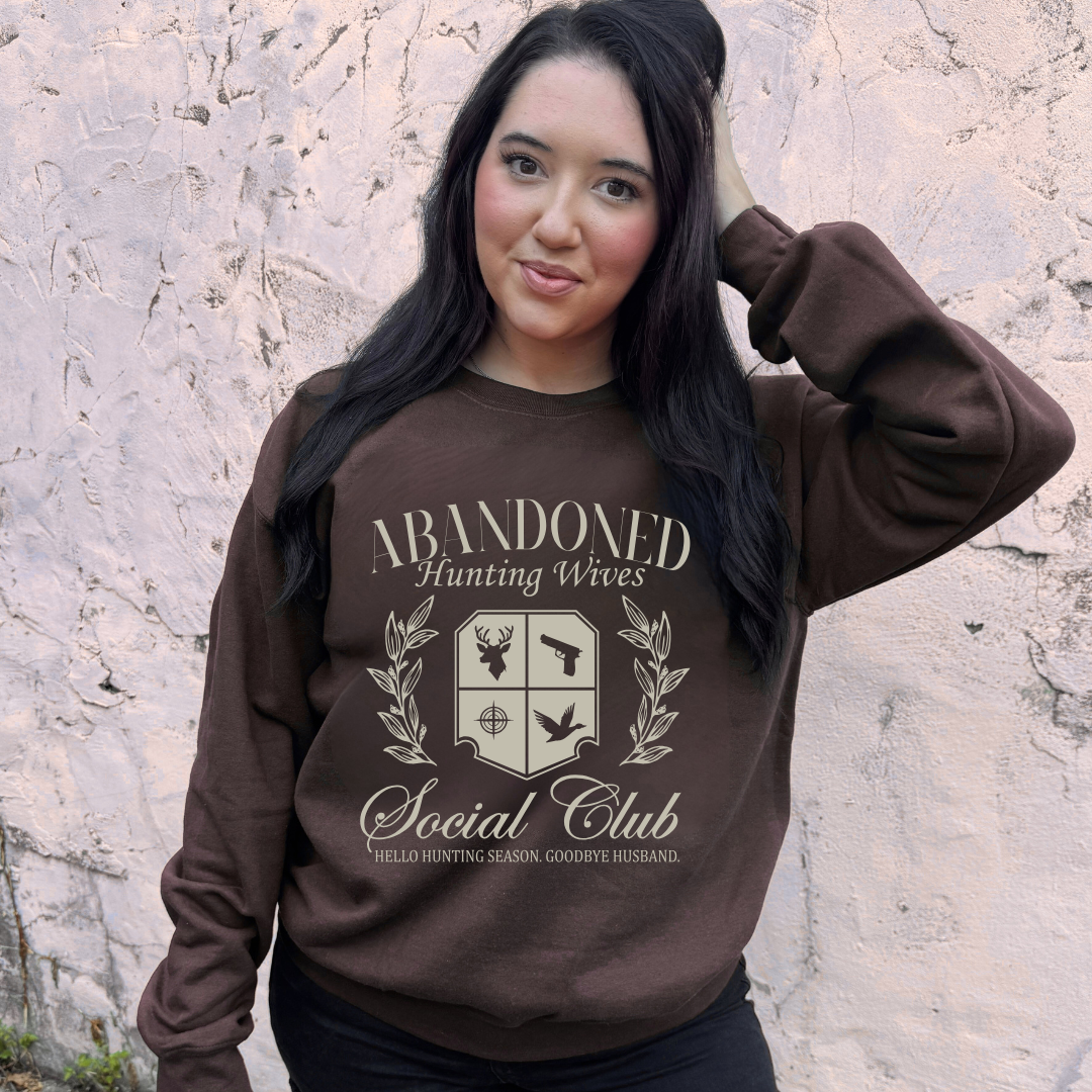 Woman wearing a brown sweatshirt with 'Abandoned Hunting Wives Social Club' design against a stone wall.