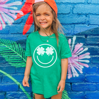 Child wearing a green shirt with a smiley face and clover design in front of a colorful mural.